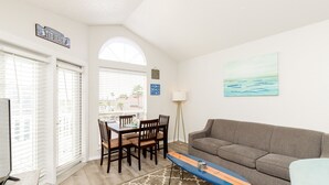 A living room with a gray sofa, a dining table with chairs, a TV, and a surfboard.