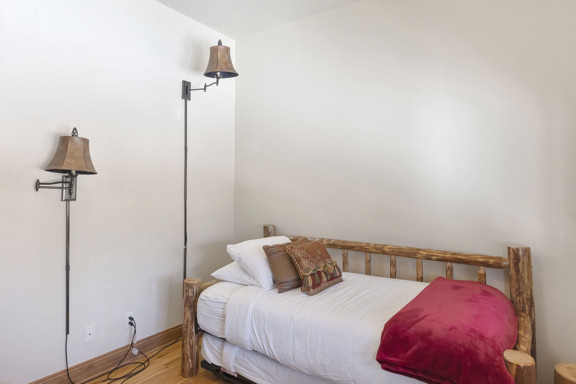A bedroom with a wooden bed, white bedding, a red throw blanket, and two wall-mounted lamps.