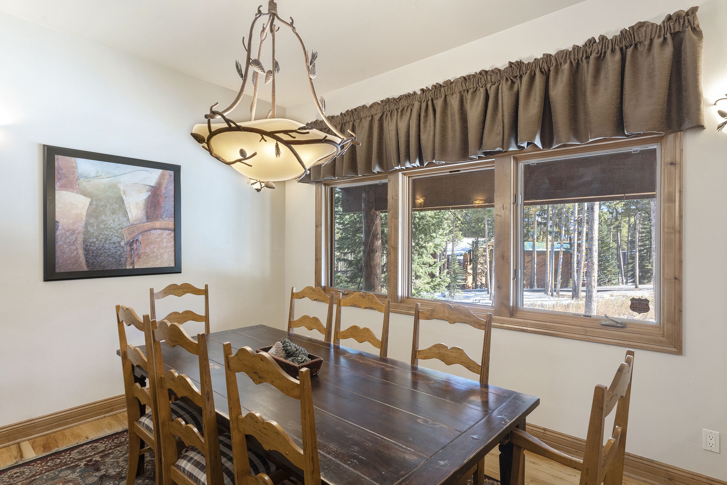 A dining area with a wooden table and chairs, a chandelier, a window with curtains, and a framed picture on the wall.