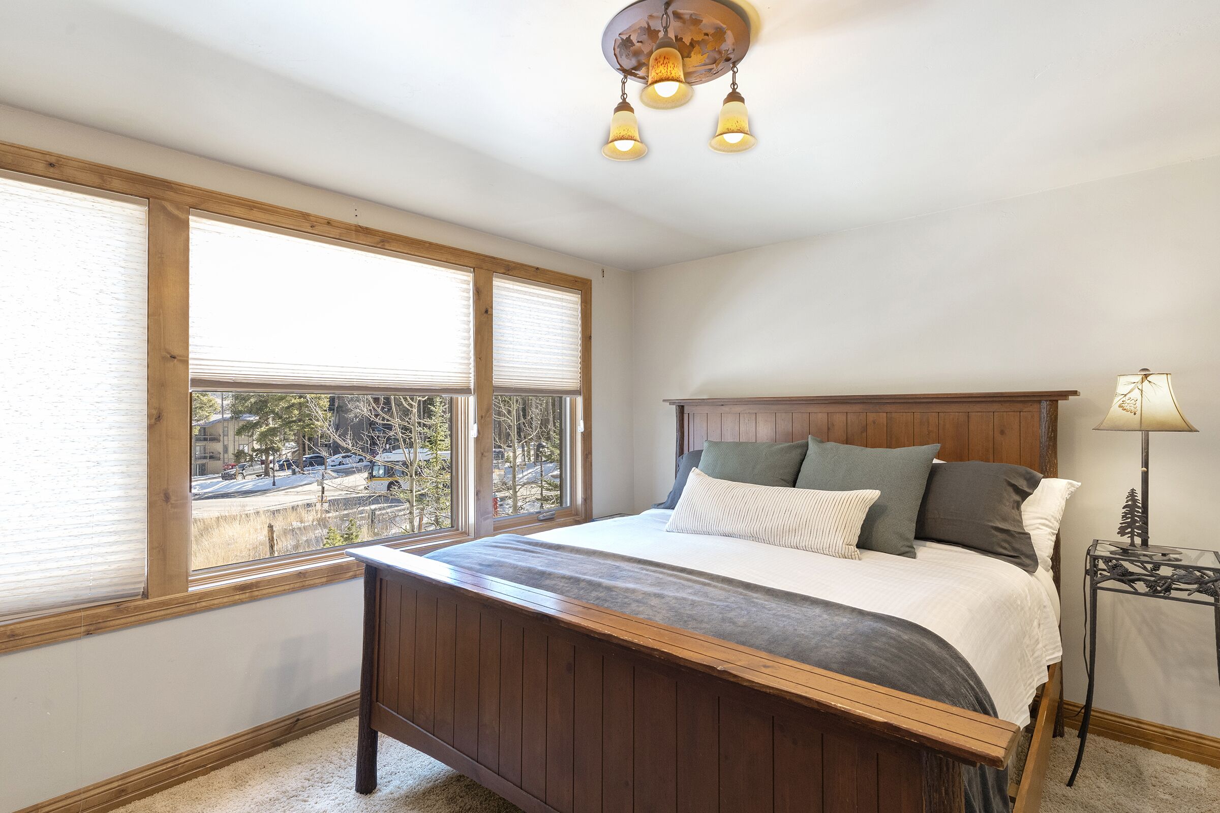 A bedroom with a wooden bed, a nightstand, a lamp, and a view of the outside through large windows.