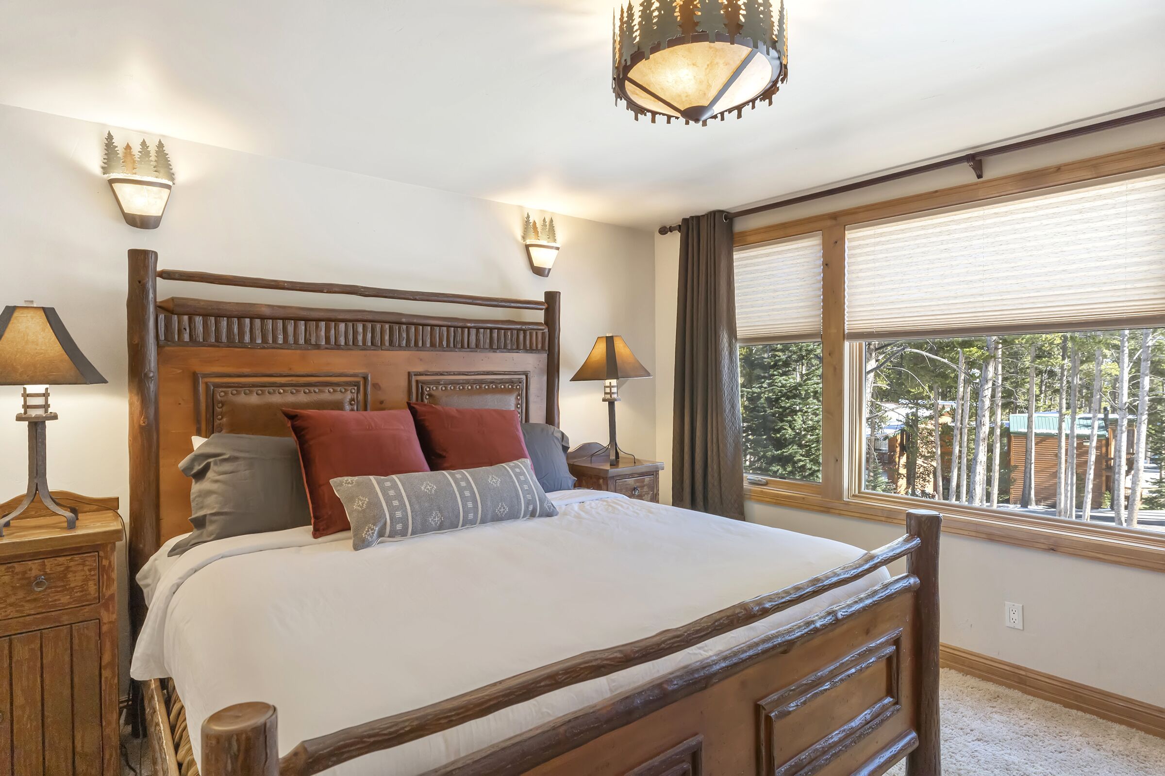 A bedroom with a wooden bed, two bedside lamps, a chandelier, and a window with blinds.