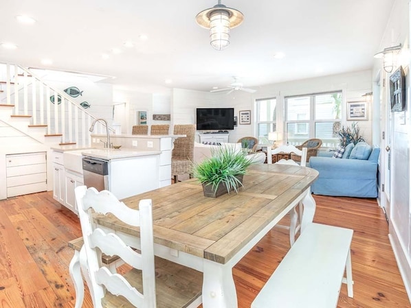 A spacious living area with a wooden dining table, white chairs, a blue sofa, and a kitchen area with white cabinets and a TV.