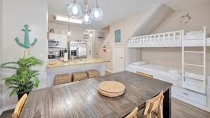 A compact, modern kitchen with a central island, a dining area with a wooden table, and a bunk bed with a white frame. - Coastal Retreat W/ Pool Views, Private Balconies and Spacious Living (Corpus Christi)