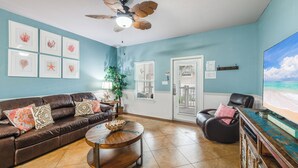 Un salon avec un canapé en cuir marron, une table basse en bois, un ventilateur de plafond, des œuvres d’art encadrées accrochées au mur et un téléviseur diffusant une scène de plage.