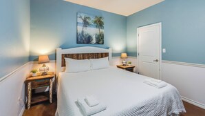 Une chambre à coucher avec une tête de lit en bois, un couvre-lit blanc, deux tables de chevet avec des lampes, un tableau représentant des palmiers et une porte blanche.