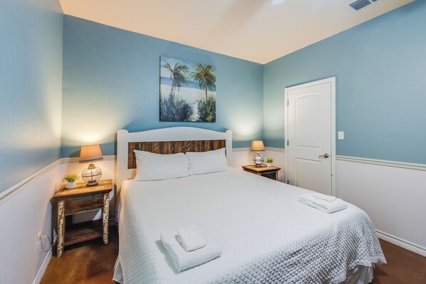 Une chambre à coucher avec une tête de lit en bois, un couvre-lit blanc, deux tables de chevet avec des lampes, un tableau représentant des palmiers et une porte blanche.