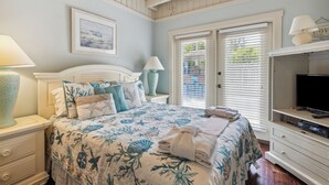 A bedroom with a bed, nightstands, a TV, and a window with blinds. - 15 Bittern Street | Cozy Home with Cottage & Pool | 4th Row | STR #028425 (Hilton Head Island)
