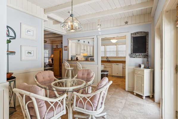 A dining area with wicker chairs and a glass table, a kitchen with white cabinets, and a large mirror above the counter.