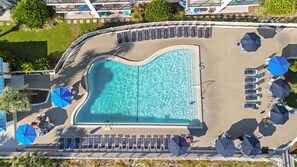 An aerial view of a swimming pool with sun loungers and umbrellas, surrounded by a white fence and greenery.