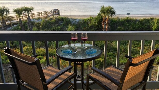 A balcony with two wicker chairs and a glass table, overlooking a beach with a ship in the distance.