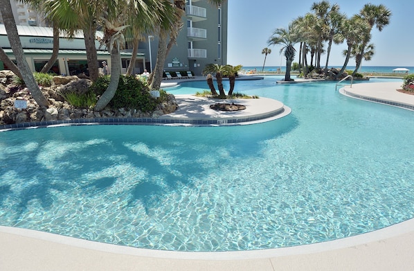 A clear blue pool with a circular island, palm trees, and a building with a sign reading "HANG OUT".