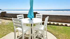 A round table with four white chairs and a blue umbrella at a beachside location.