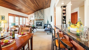 A spacious living area with a stone fireplace, wooden ceiling, and a kitchen area with bar stools and a fruit bowl.