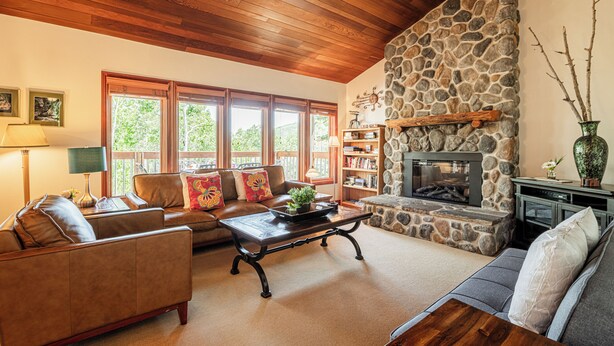 A cozy living room with a stone fireplace, wooden ceiling, and large windows.