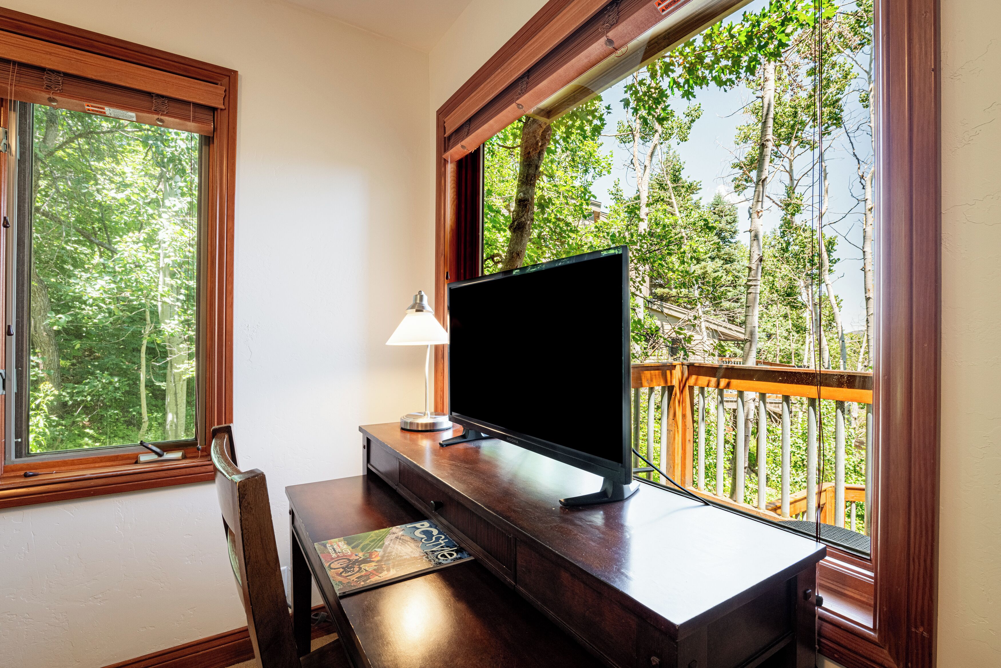 Une pièce comprenant un bureau, une chaise, un téléviseur et une fenêtre donnant sur des arbres et une terrasse en bois.