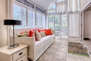 Una habitación acogedora con un banco blanco, una lámpara y vistas amplias a través de ventanas con contraventanas.