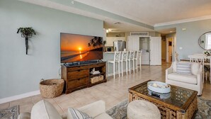 A living room with a TV, white chairs, a sofa, and a kitchen in the background. - 2403 SeaCrest-4th Floor/ Ocean Views & Newly Updated (Hilton Head Island)