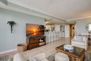 A living room with a TV, white chairs, a sofa, and a kitchen in the background.