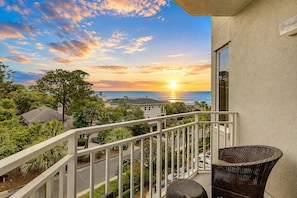 A balcony with a view of the sunset over the ocean, surrounded by greenery and residential buildings.