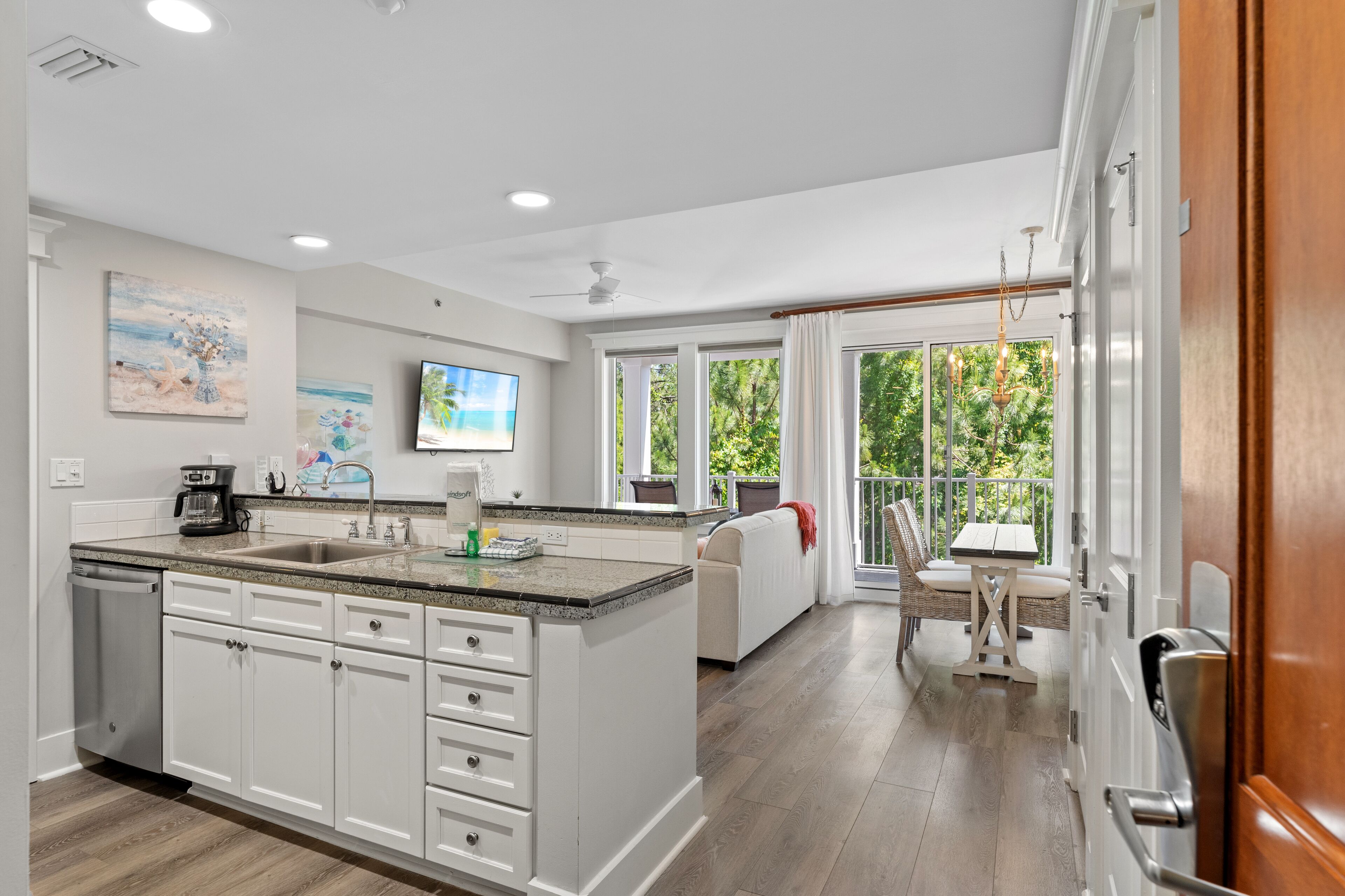 A modern kitchen with white cabinets, granite countertops, and a large island. There is a dining area with a table and chairs outside, and a view of greenery through large windows.