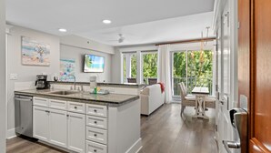 A modern kitchen with white cabinets, granite countertops, and a large island. There is a dining area with a table and chairs outside, and a view of greenery through large windows.