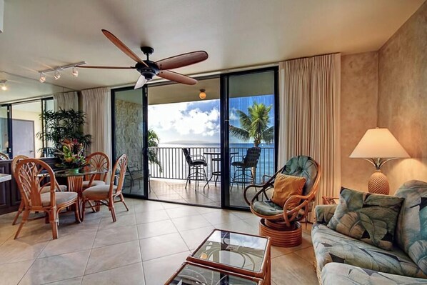 A spacious living area with a balcony, wicker furniture, a ceiling fan, and a view of the ocean.