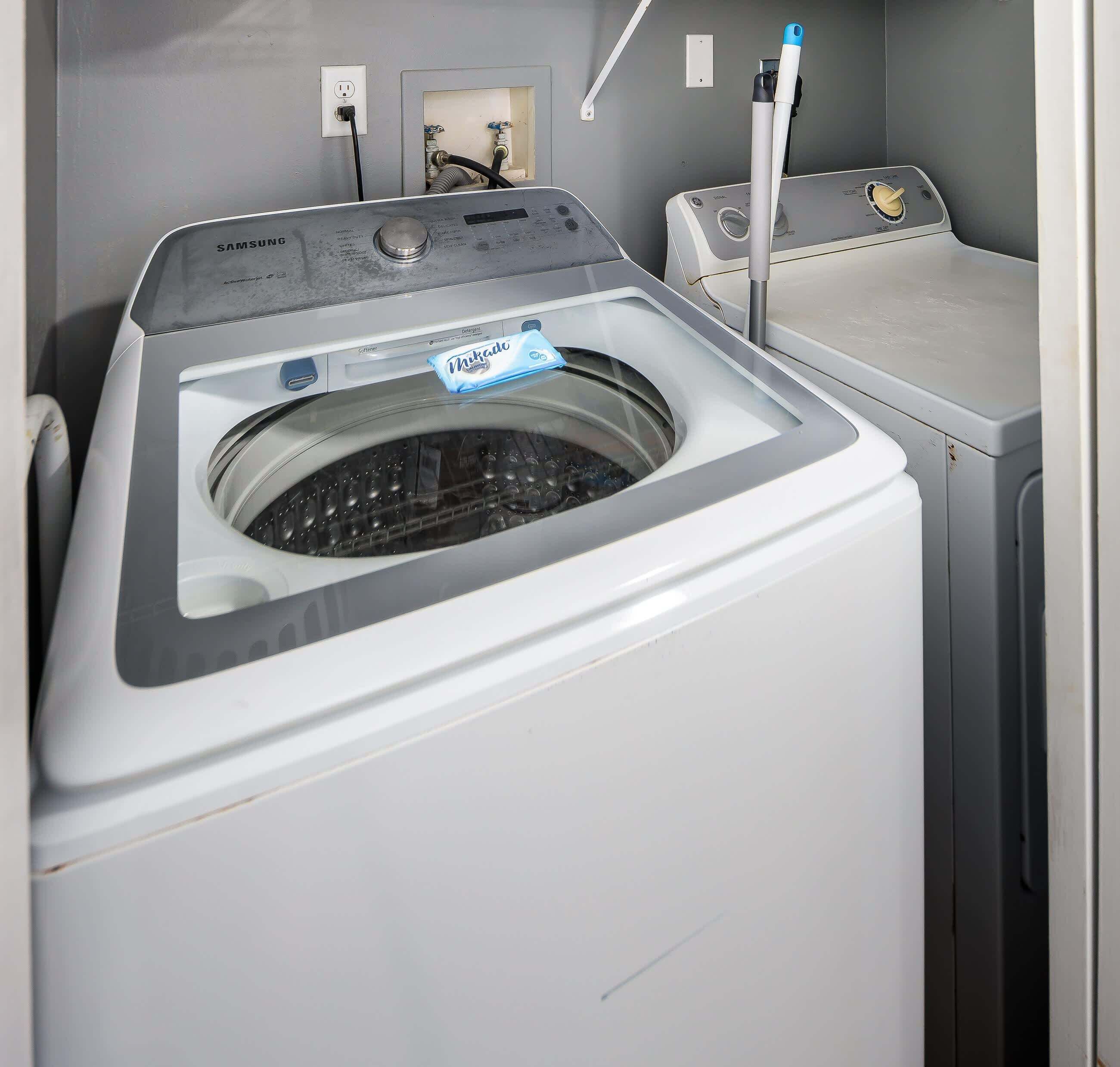 A front-loading washing machine with a control panel, a power outlet, and a water inlet valve on the wall.