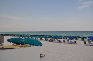 A beach with rows of blue umbrellas and lounge chairs, a sailboat in the distance, and a clear sky.