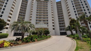 Un hôtel à plusieurs étages, doté d’une entrée principale et entouré de jardins aménagés et de palmiers.