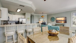 A modern kitchen with white cabinets, a stainless steel refrigerator, and a dining area with wicker chairs and a glass table. - 1106 SeaCrest-Oceanviews, walk to dining, shopping & activities. (Hilton Head Island)