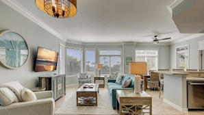 A spacious living room with a large clock, a television, and a view of the ocean through the windows. - 1106 SeaCrest-Oceanviews, walk to dining, shopping & activities. (Hilton Head Island)