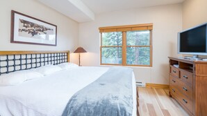 Une chambre à coucher comprenant un lit, un téléviseur, une commode, une fenêtre donnant sur des arbres et un tableau encadré accroché au mur.