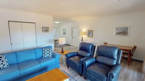 A living room with a blue sofa, two leather armchairs, a wooden coffee table, and a dining area with a wooden table and chairs. - Atlantic Breeze #7 2 bedrooms, 1 bathroom (Jekyll Island)