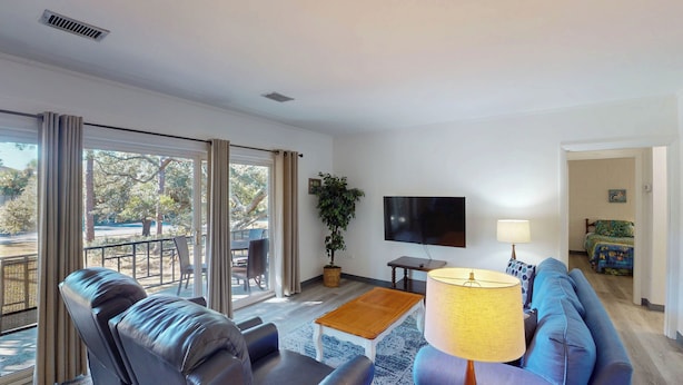 A living room with a sofa, a coffee table, a TV, and a lamp. There is a balcony with a view of trees and a street. - Atlantic Breeze #7 2 bedrooms, 1 bathroom (Jekyll Island)
