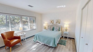A bedroom with a bed, two bedside tables with lamps, a chair, and a window with blinds. - Atlantic Breeze #7 2 bedrooms, 1 bathroom (Jekyll Island)