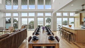 A spacious dining area with a long wooden table set for a meal, large windows offering ocean views, and a modern kitchen adjacent to the dining space. - Magnolia: 7 BR Beachfront Home - Pet Friendly, Priv. Pool, Sleeps 24! (Cape San Blas)