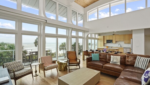 A spacious living area with a large window overlooking the ocean, a cozy seating arrangement, and a modern kitchen in the background. - Magnolia: 7 BR Beachfront Home - Pet Friendly, Priv. Pool, Sleeps 24! (Cape San Blas)