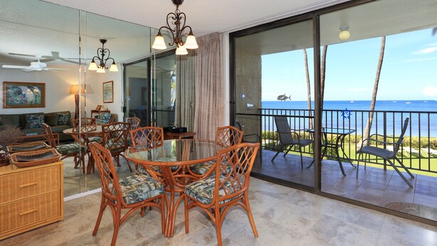 A dining area with wicker chairs and a glass table, a ceiling fan, and a view of the ocean through large glass doors. - Unwind by the Waves: Kihei Beach #203, Prime Beachfront Tropical Escape! (Kihei, Maui)