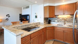A kitchen with granite countertops, wooden cabinets, a stainless steel refrigerator, and a built-in stove. - Unwind by the Waves: Kihei Beach #203, Prime Beachfront Tropical Escape! (Kihei, Maui)