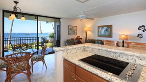 A kitchen with a large island, a ceiling fan, and a view of the ocean through a sliding glass door. - Unwind by the Waves: Kihei Beach #203, Prime Beachfront Tropical Escape! (Kihei, Maui)