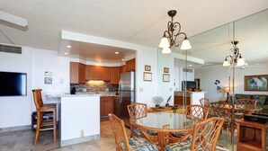 A modern kitchen with wooden cabinets, a stainless steel refrigerator, and a dining area with wicker chairs and a glass table. - Unwind by the Waves: Kihei Beach #203, Prime Beachfront Tropical Escape! (Kihei, Maui)