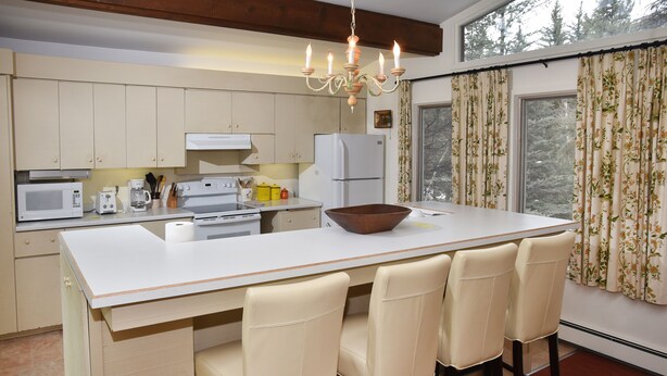 Una cocina con una isla central, armarios blancos, un microondas, una estufa y un refrigerador. Hay cuatro sillas alrededor de la isla y una araña colgante arriba.