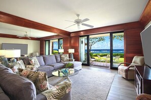 A modern living room with a large sofa, a coffee table, and a view of the ocean through large glass doors. - Poipu Kapili 10 Ocean Front By Suite Paradise (Koloa)