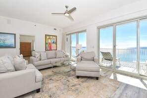 Relax in this open-plan living space with a sofa, armchairs, and a glass coffee table, offering a view of the ocean through floor-to-ceiling glass doors.