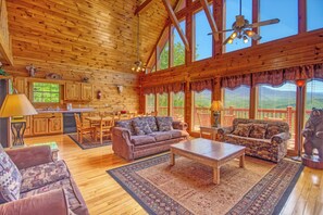 A spacious cabin interior with a high ceiling, wooden walls, a dining area, and a large window offering a view of the outdoors. - Cabin w/Amazing Mountain Views-Hot Tub-Game Room (GATLINBURG)