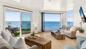 Ein geräumiges Wohnzimmer mit einem großen Schiebeglasfenster, das auf einen Balkon mit Meerblick führt. Es gibt ein Sofa, einen Couchtisch und einen an der Wand befestigten Fernseher.