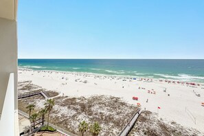 Vista a la playa desde un balcón con terraza de madera y palmeras, una playa arenosa con sombrillas y un cielo azul despejado.