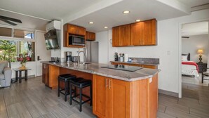 A modern kitchen with wooden cabinets, a central island, and a dining area with a table and chairs. - Oceanview Oasis Walk to Shops & Dining: Kamaole Nalu #303 Convenience & Charm! (Kihei)