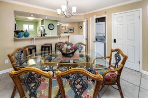 A dining area with a glass-top table surrounded by ornate chairs, a chandelier, and a kitchen in the background.