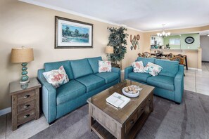 A living room with two teal sofas, a wooden coffee table, and a side table with a lamp. A framed picture hangs on the wall, and a plant is visible in the background.